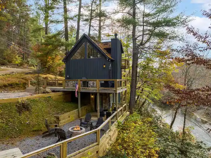 Off-season riverside cabin rental: black A-frame amid autumn trees with empty fire pit seating by a quiet riverbank