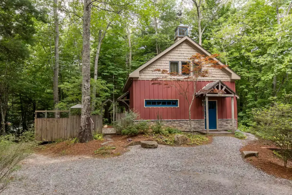 Rustic red forest cabin surrounded by green trees and empty gravel driveway, offering a quiet off-season retreat vibe