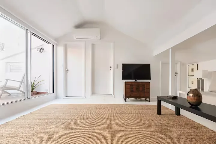 Bright minimalist vacation rental living room with woven rug, low bench and TV opening to empty balcony in off-season calm
