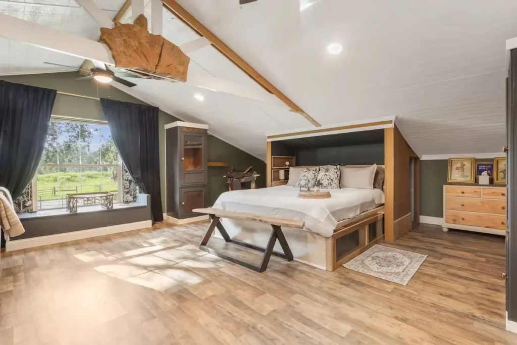 Cozy loft bedroom with rustic king bed and large window overlooking quiet green meadow in off-season retreat.