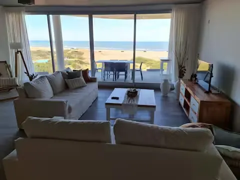 Bright beachfront living room with cozy sofas, empty patio and calm off-season beach view through floor-to-ceiling windows.