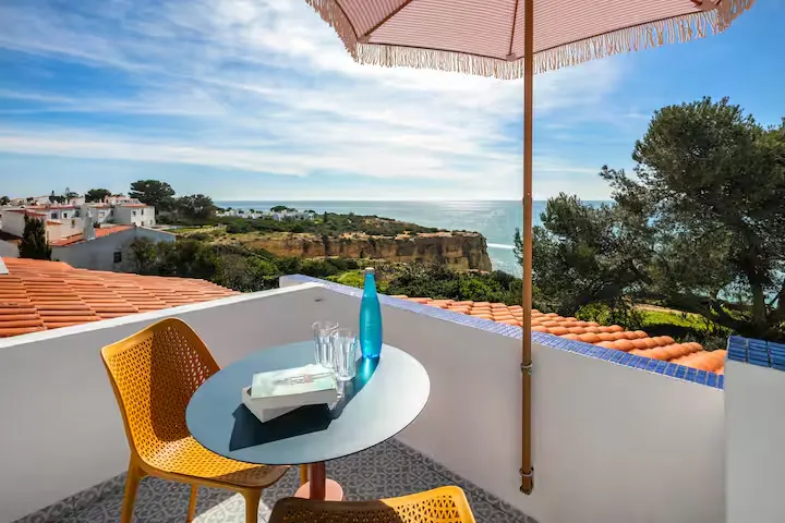 Sunlit off-season terrace with coastal cliff and calm sea view, empty chairs, table, umbrella, book and water bottle.