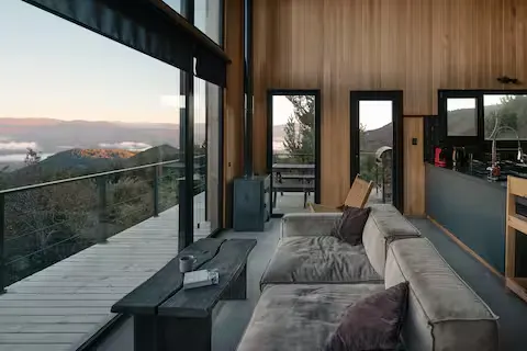 Secluded off-season mountain cabin interior with panoramic glass walls, cozy sofa and deck overlooking misty valley at dawn.