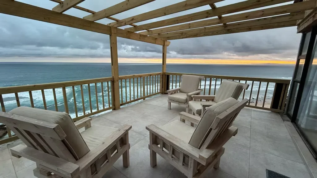 Off-season oceanfront terrace with wooden lounge chairs under a pergola, overcast sky and sunset glow over empty beach