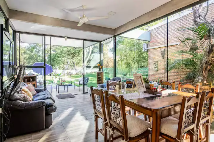 Quiet off-season travel retreat patio with glass walls, wooden dining table, sofa and lush garden view
