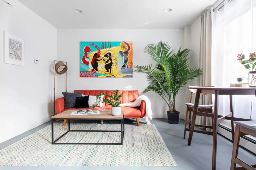 Bright off-season rental living room with orange sofa, abstract wall art, potted palm, wooden coffee table and sunlit dining nook.
