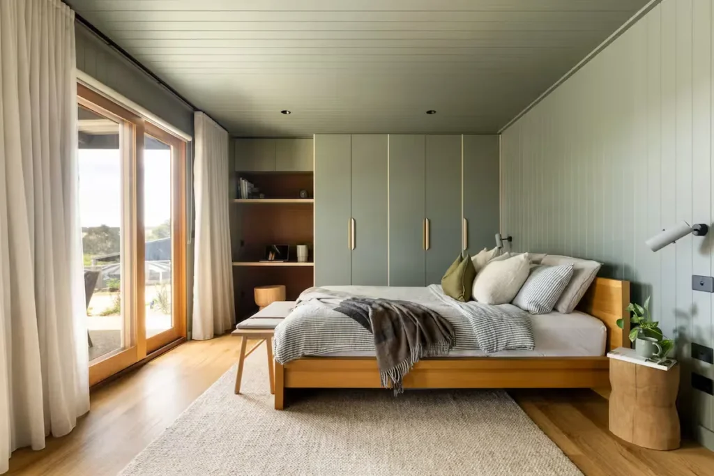 Cozy lodge bedroom with wood-frame bed, soft green paneled walls and sliding glass doors overlooking a quiet off-season patio.