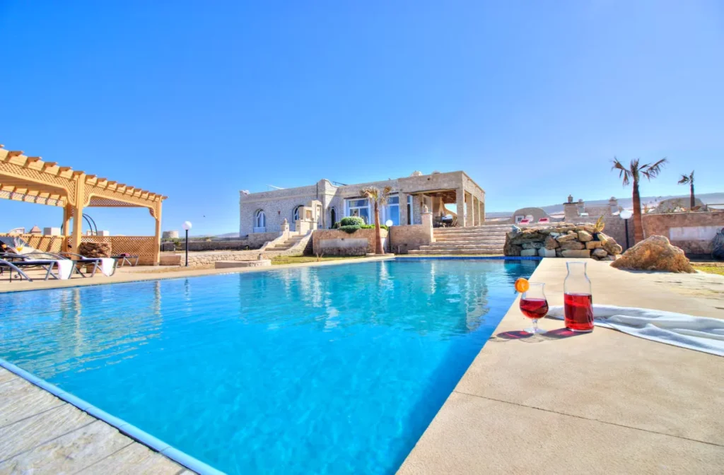 Empty poolside at a stone villa under clear off-season skies, tranquil travel retreat with sun loungers and red wine.