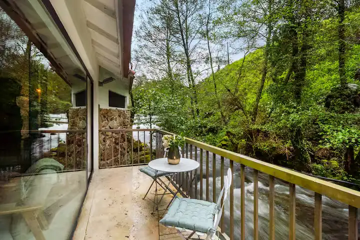 Off-season riverside cabin balcony with table overlooking rushing stream through misty green woods in peaceful retreat.