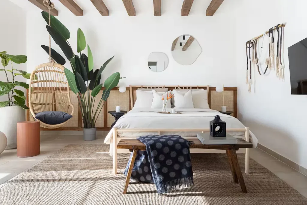 Sunlit bohemian hotel bedroom with rattan headboard, hanging wicker chair and potted plants, quiet off-season retreat.