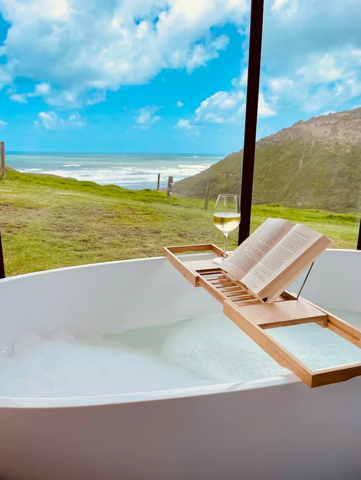 Serene off-season coastal retreat bath with book and wine, waves crashing on a grassy cliff under a bright cloudy sky