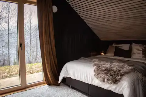 Cozy off-season cabin bedroom with fur throw, dark wood slanted ceiling and glass doors opening to a misty, leafless forest.