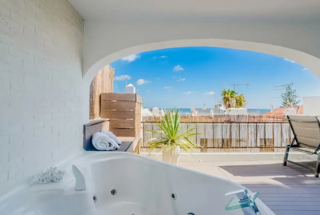 Off-season jacuzzi on a quiet sunlit terrace overlooking white rooftops and a calm blue sea under clear skies.