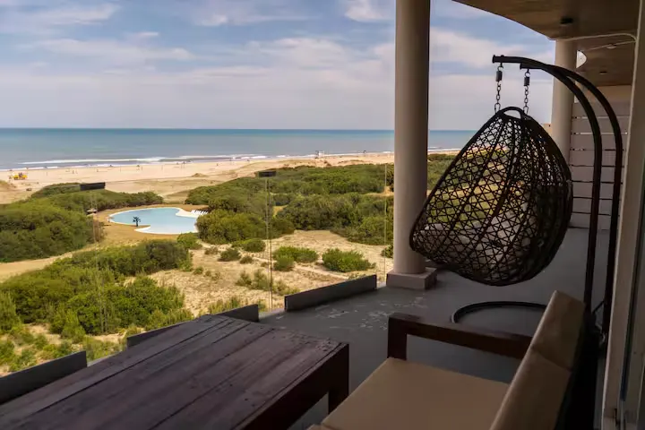 Empty vacation balcony with wicker chair overlooking quiet off-season beach, dunes and unused pool under cloudy sky.