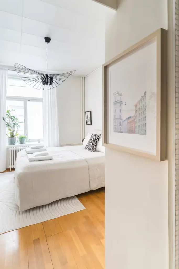 Cozy minimalist vacation apartment bedroom in off-season with bright white linens, indoor plants and warm wooden floors.