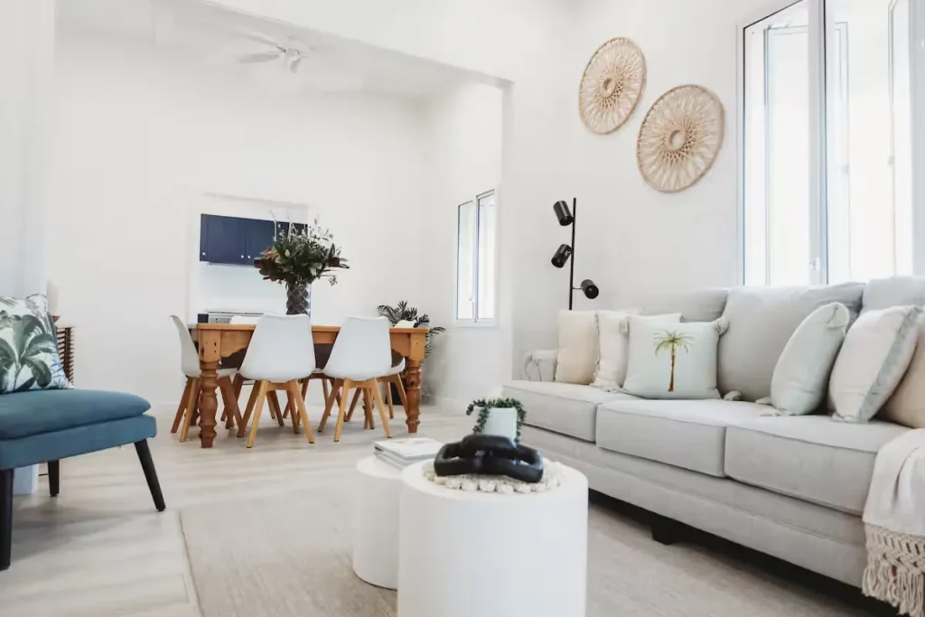 Sunlit seaside vacation rental living room with palm-print pillows and rustic wooden dining table, peaceful off-season quiet.
