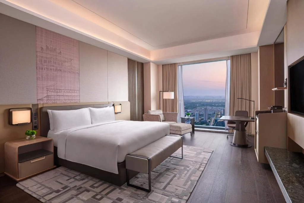 Spacious luxury hotel room with king bed and city skyline view at dusk, offering peaceful off-season serenity.
