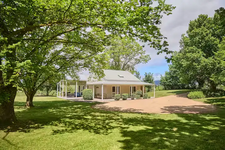 Serene country cottage with wraparound porch nestled under large trees, lush green lawn and tranquil off-season travel vibes
