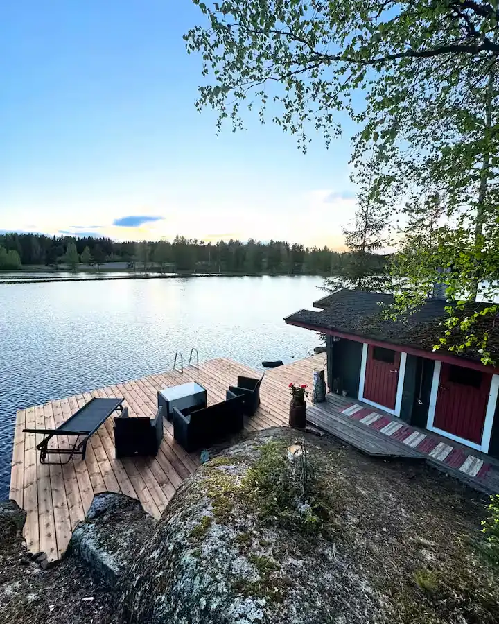 Serene off-season lakeside deck with lounge seating by a rustic red boathouse at sunset, quiet forest shoreline beyond.
