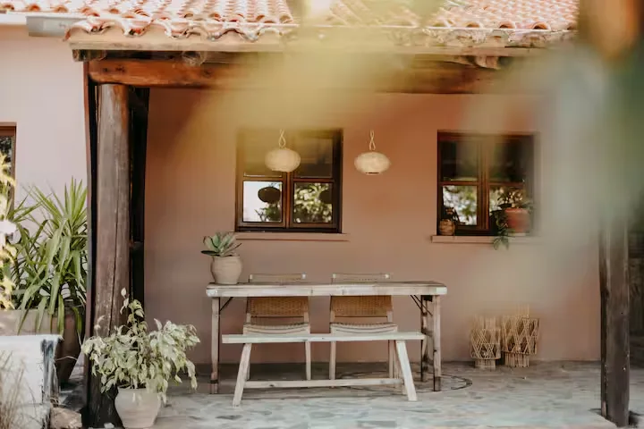 Quiet sunlit terrace travel scene with empty wooden bench and table under terracotta roof, potted plants and off-season vibe