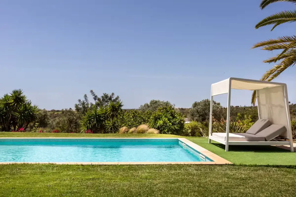 Empty poolside daybed beside a turquoise pool under clear sky, surrounded by lush greenery for serene off-season relaxation.