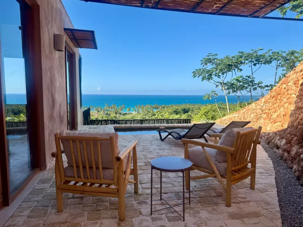 Empty thatched-roof tropical villa terrace overlooking turquoise ocean and palm trees in serene off-season calm.