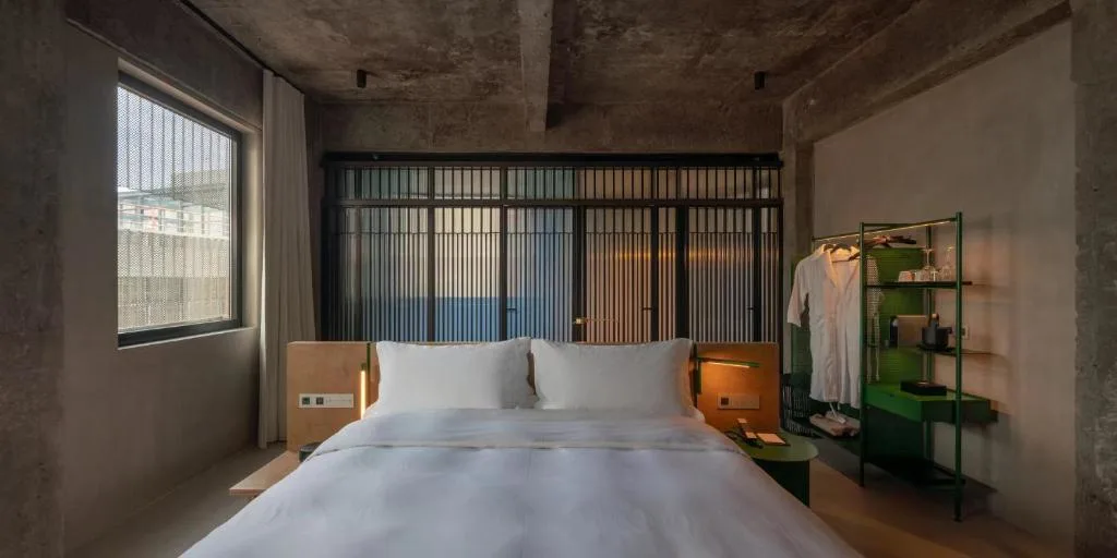 Minimal industrial boutique hotel room with king-size bed and open wardrobe, soft off-season light from large window.