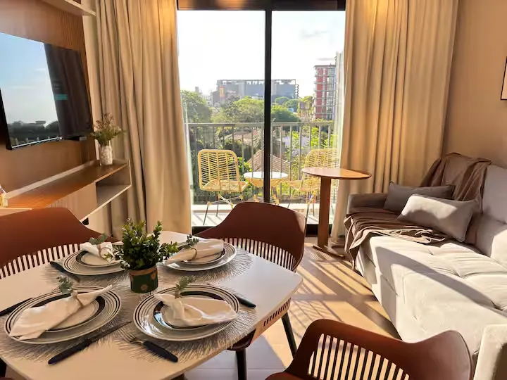 Sunlit off-season vacation rental with dining table set for four and empty balcony chairs overlooking a quiet city.