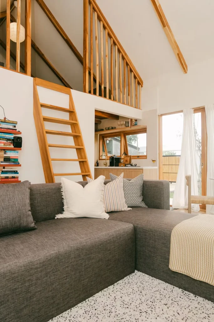 Cozy off-season travel cabin retreat: living area with grey sofa, decorative pillows, wooden loft ladder and natural light