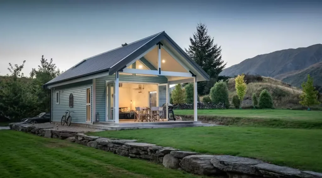 Cozy modern mountain cabin at dusk with warm patio lights, empty lawn and rolling hills evoking a serene off-season getaway.
