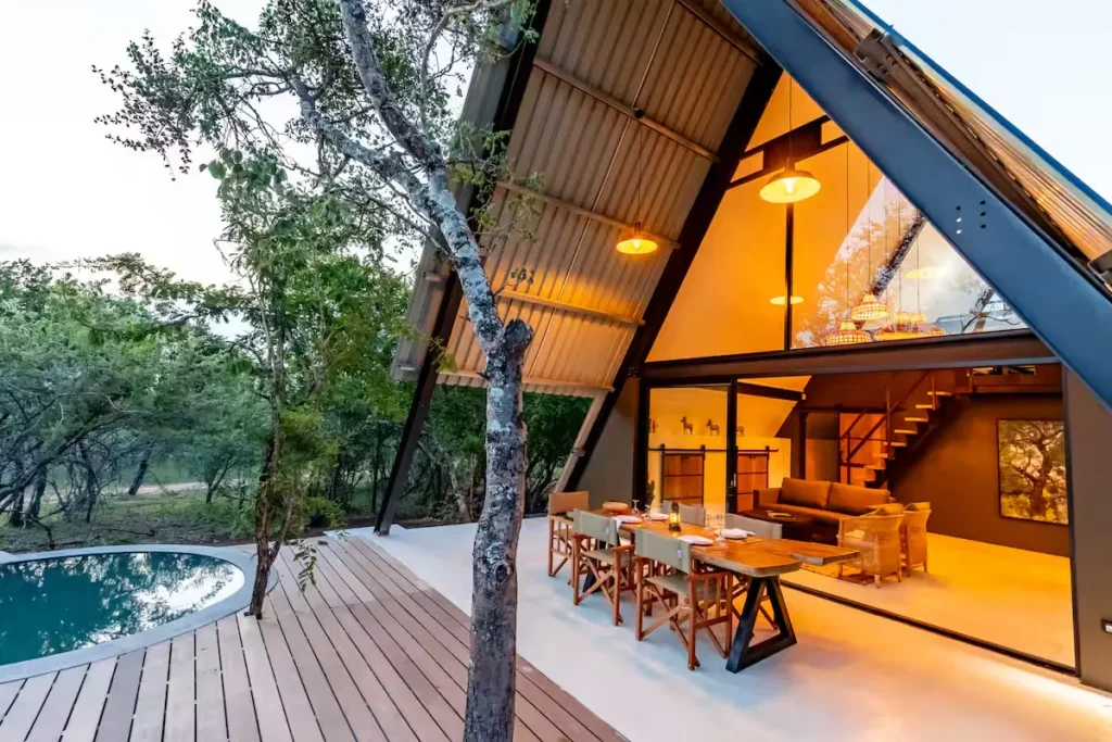 Cozy luxury A-frame cabin retreat in quiet woods at dusk, glowing interior, empty deck and pool for off-season travelers.