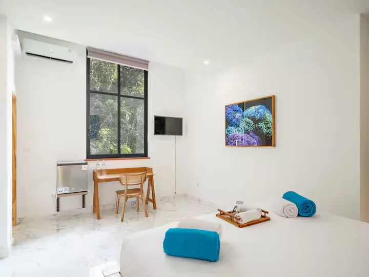 Minimalist bright hotel room with desk, TV and bed with rolled towels by a tropical view, evoking off-season calm.