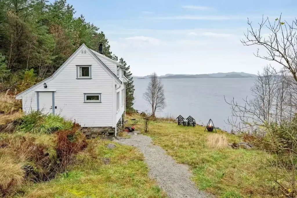 Secluded white lakeside cabin with chairs by calm water in off-season, autumn grass and bare trees evoke tranquil retreat