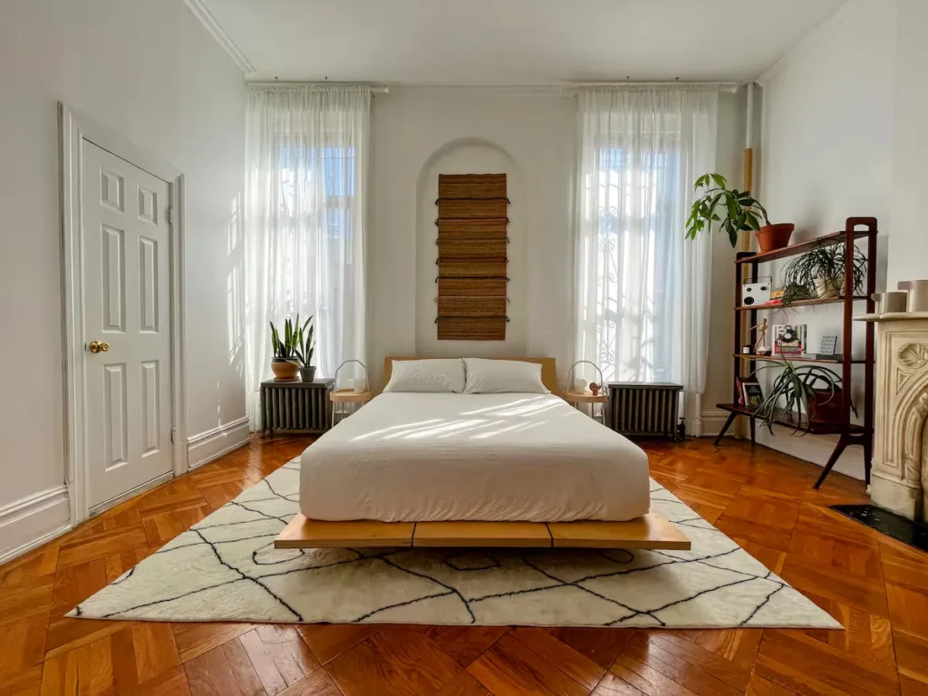 Sunlit minimalist bedroom with platform bed, wooden floors and potted plants, offering a serene off-season hotel retreat.