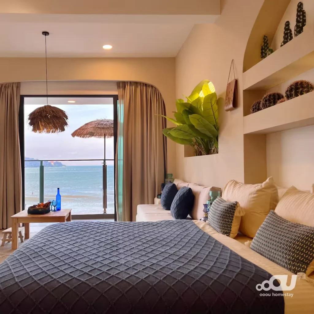 Cozy seaside homestay bedroom with pillows and ocean view balcony overlooking empty off-season beach and thatched umbrellas.
