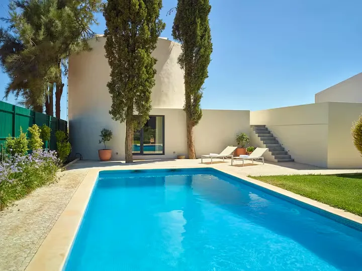 Off-season vacation villa scene: empty private pool, sun loungers and tall cypress trees beneath a clear blue sky.