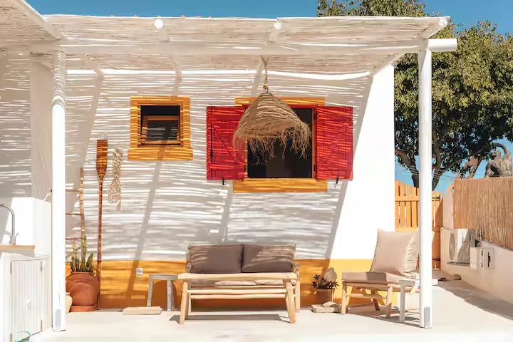 Quiet off-season seaside terrace with wicker seating, pergola-shadowed walls and open red shutters under a sunny sky