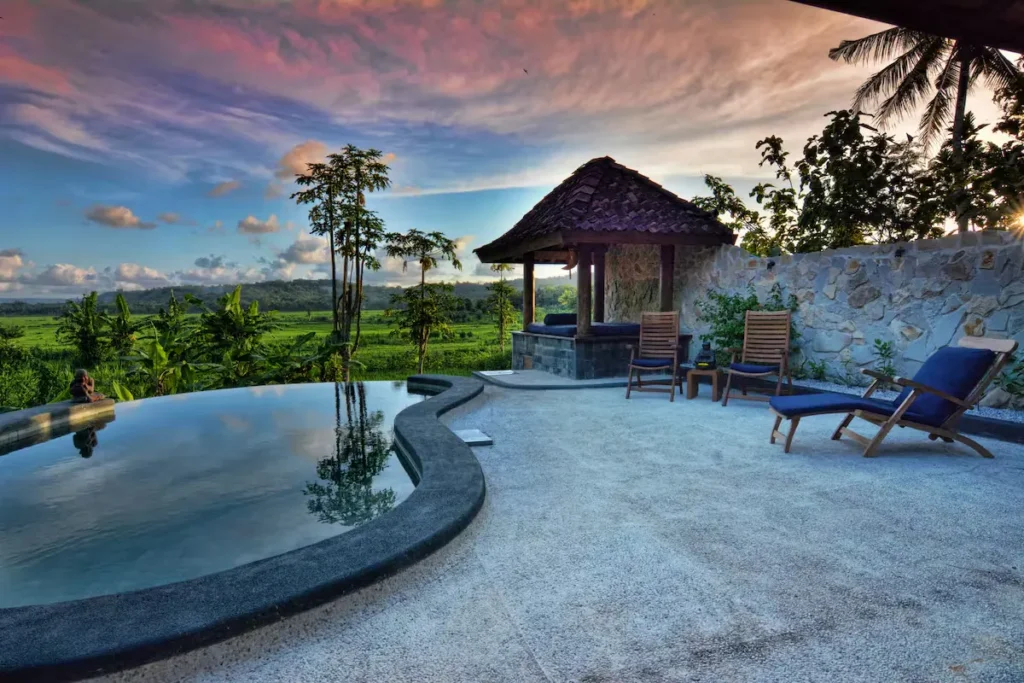 Off-season infinity pool deck at sunset, empty lounge chairs beside a gazebo, overlooking lush green fields under pastel sky