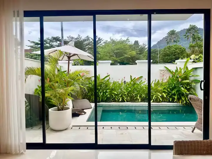 Looking through glass doors at a quiet off-season tropical villa patio with private pool, lush greenery, empty lounge chairs.