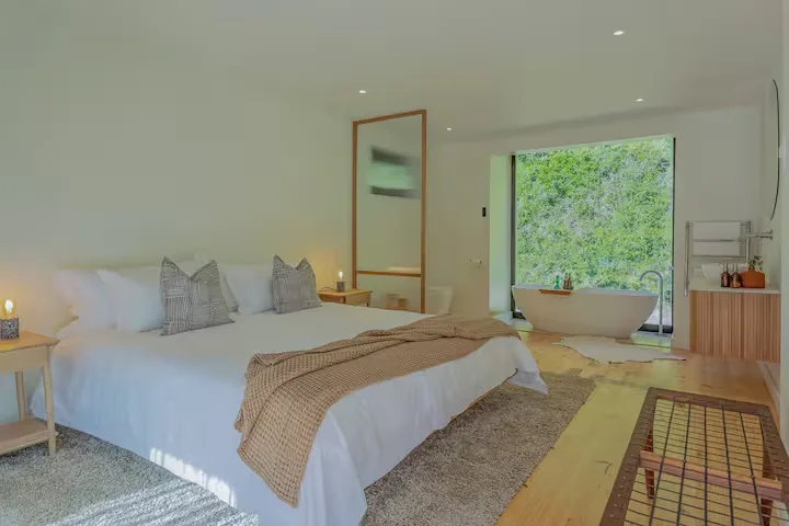 Empty modern hotel suite with king bed, bathtub by open window overlooking lush greenery in off-season stillness