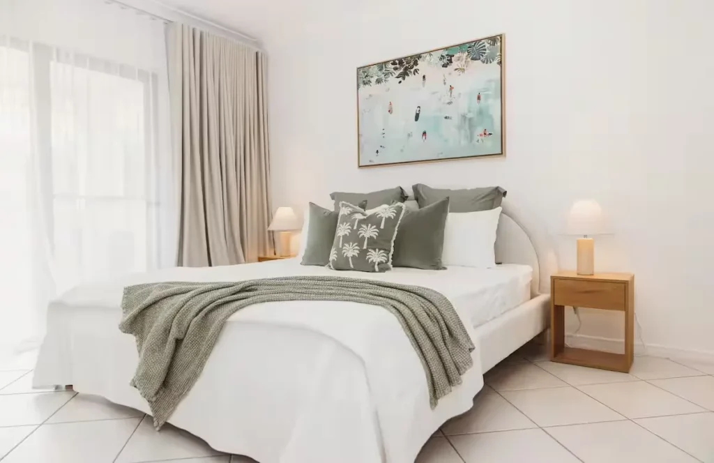 Bright empty vacation rental bedroom with white linens and sage-green pillows, quiet off-season seaside getaway feel.