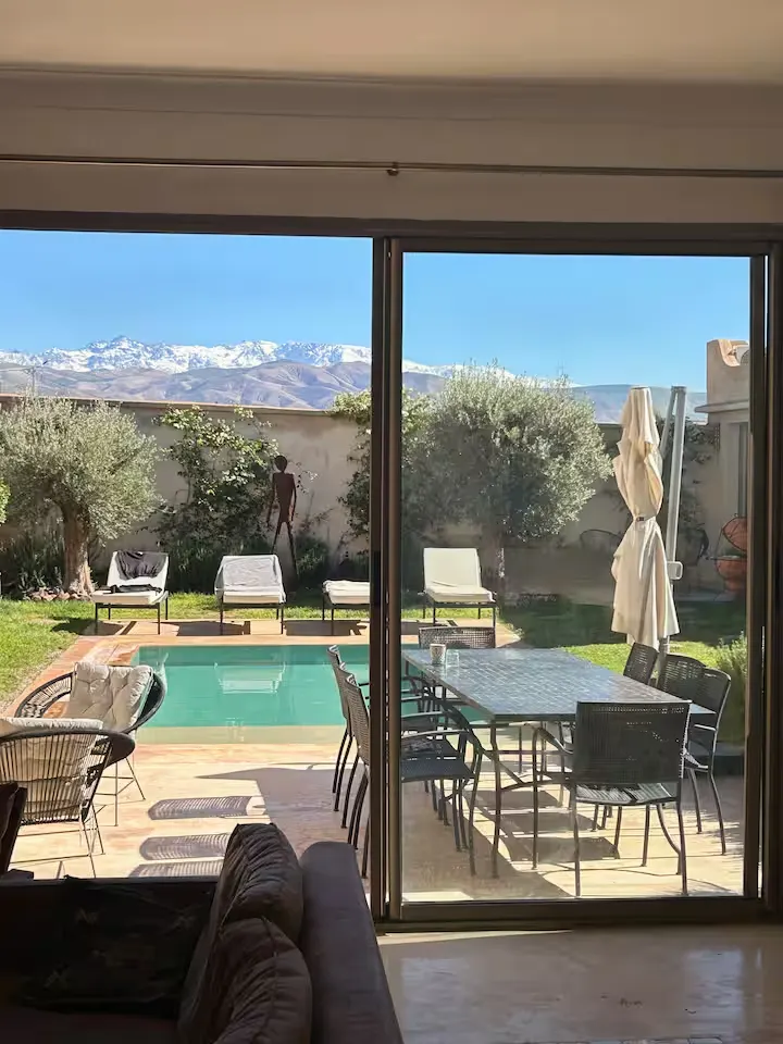 Quiet off-season villa view: private pool, lounge chairs, olive trees and snow-capped peaks through sliding glass doors.
