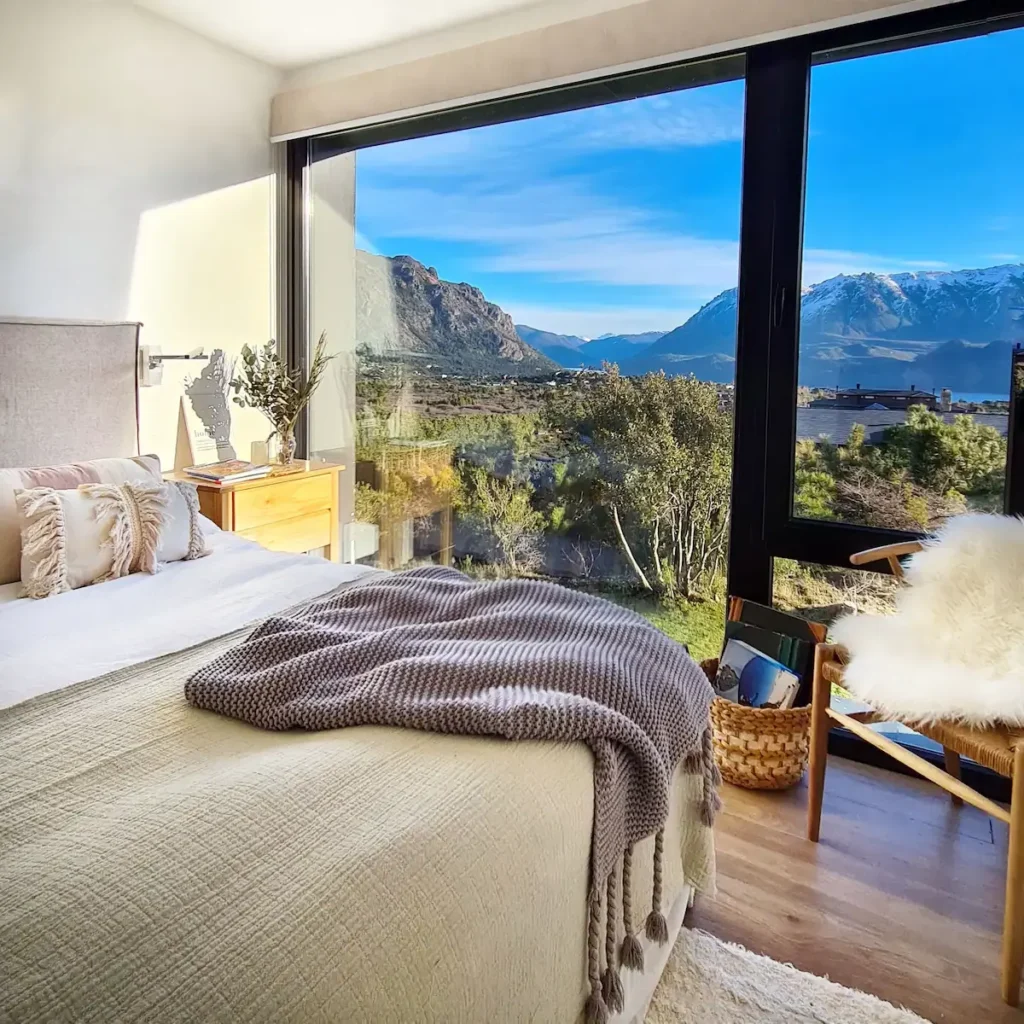 Cozy lakeside mountain cabin bedroom with panoramic glass view of snow-capped peaks and tranquil off-season landscape.
