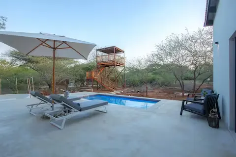 Off-season poolside loungers under umbrella beside a quiet safari viewing tower amid dry-season savanna shrubs.