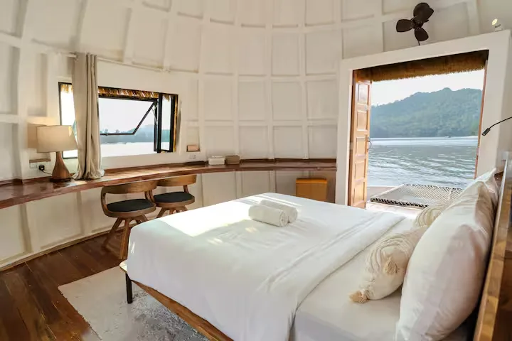 Empty off-season lakeside bungalow with cozy white bedding, wooden desk and open doors to tranquil mountain water view.