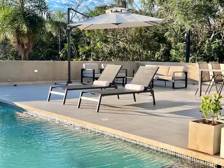 Quiet sunlit poolside terrace in off-season, empty lounge chairs under umbrella with lush tropical greenery backdrop.