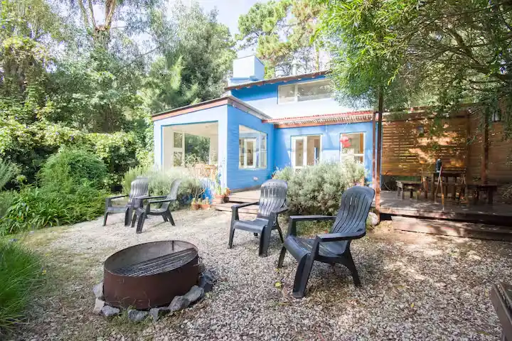 Quiet off-season retreat: blue woodland cottage patio with empty chairs around a fire pit under late autumn trees.
