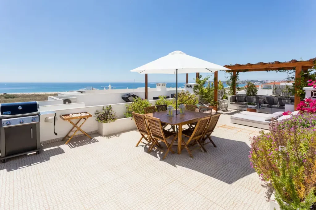 Quiet off-season seaside rooftop terrace with wooden dining table, umbrella, ocean views and sunny coastal ambiance.