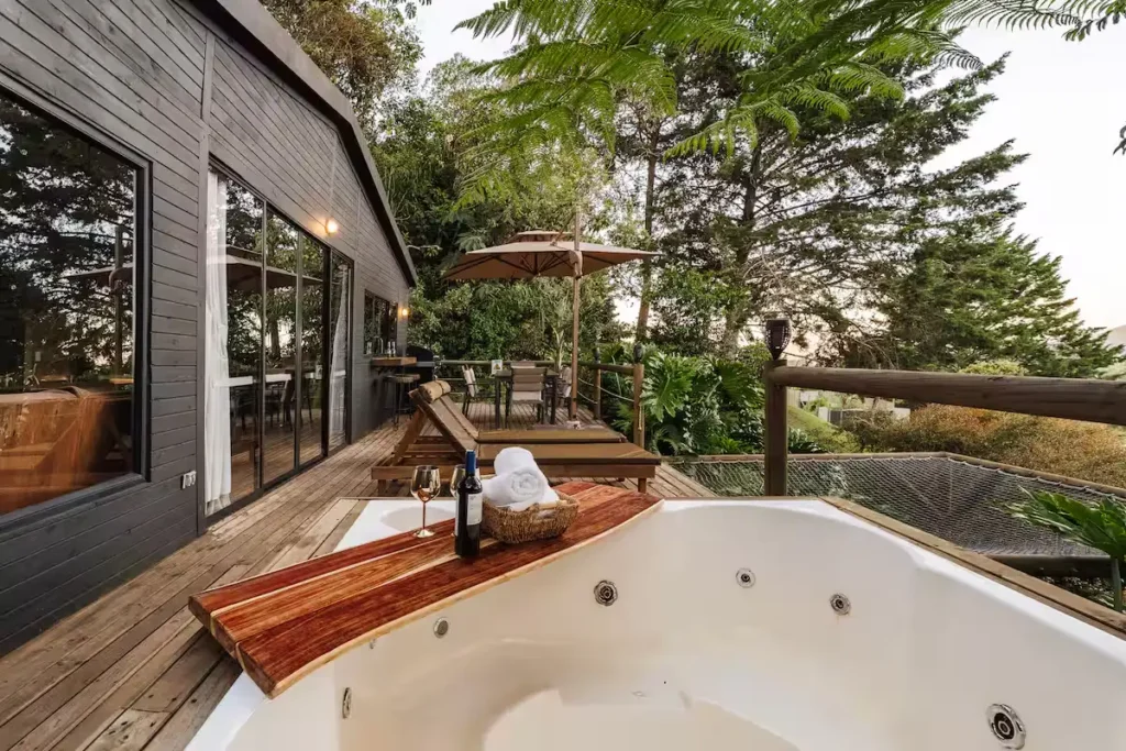 Luxury cabin deck with empty hot tub, wine bottle and glass, loungers under umbrella among lush trees in off-season retreat