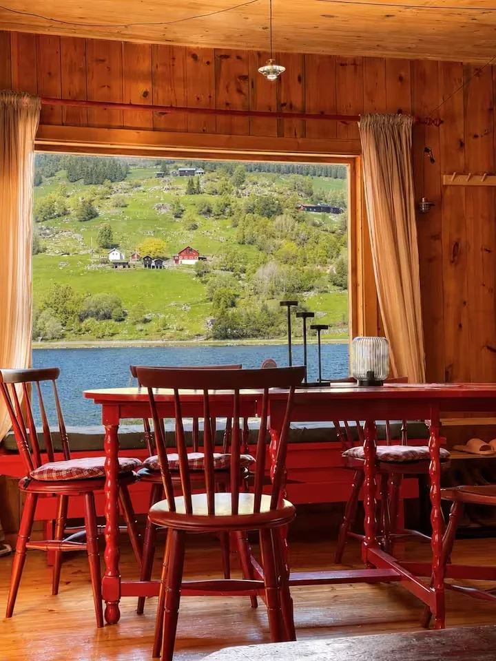 Cozy lakeside cabin interior with red wooden chairs and table overlooking a tranquil off-season lake and green hillside view